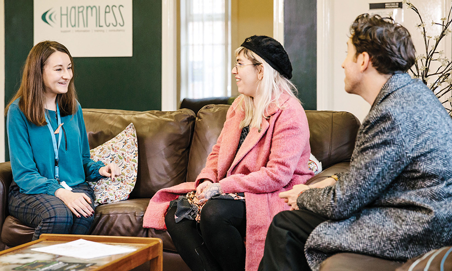 Three people sit on a brown sofa in a cosy room, engaged in a friendly conversation.