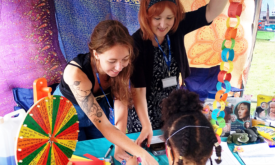 Two women and a child smile making colourful crafts outdoors, with paper chains and a bright spinner.