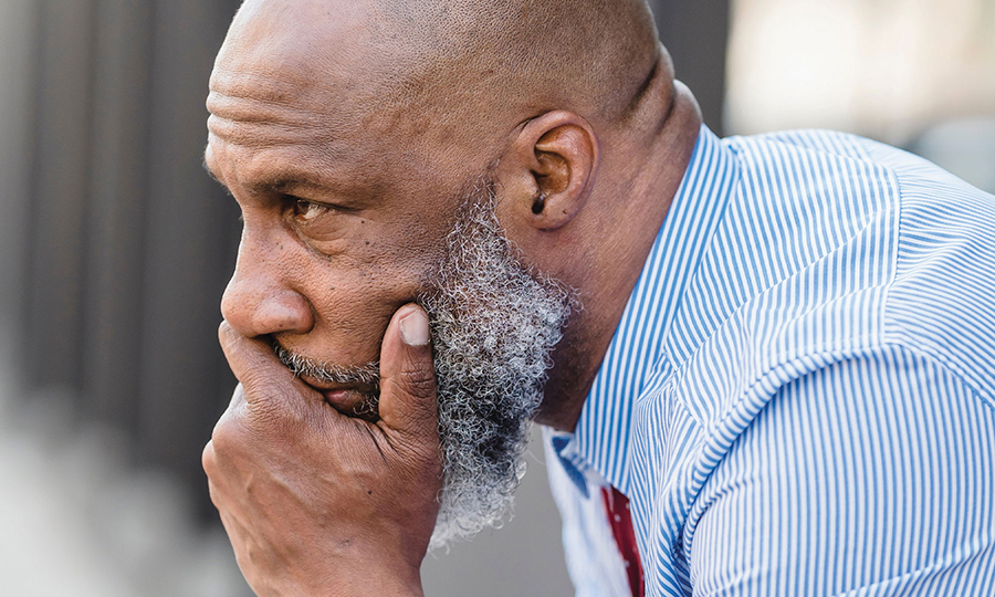 An older man with a beard, wearing a striped shirt, deep in thought, rests his chin on his hand.