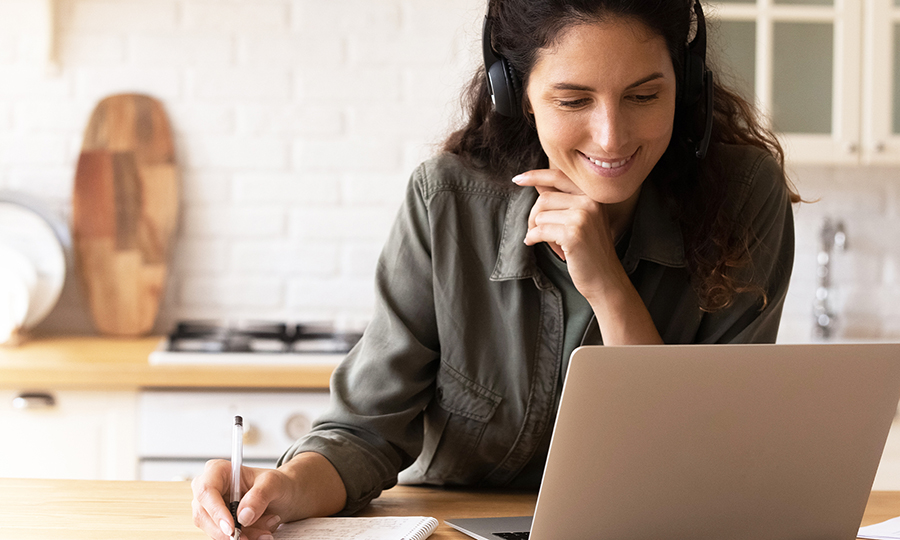A woman wearing headphones smiles while working on a laptop in a bright kitchen.