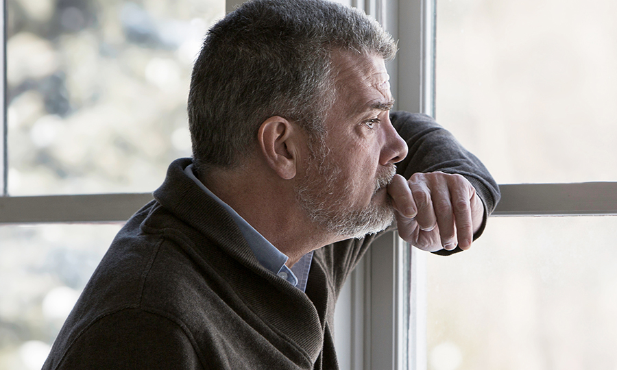 A mature man with a grey beard gazes pensively out a window, resting his chin on his hand.
