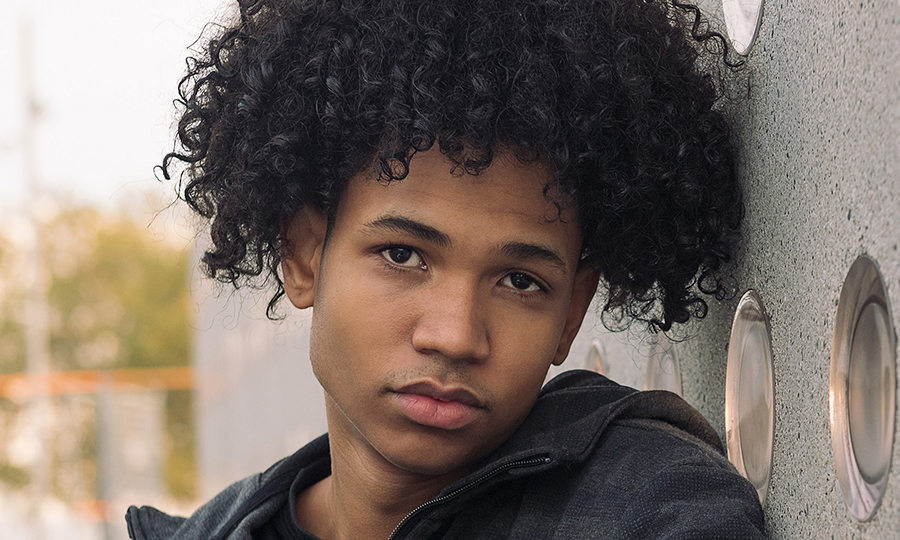 A young person with curly hair leans against a textured wall, looking pensively at the camera.