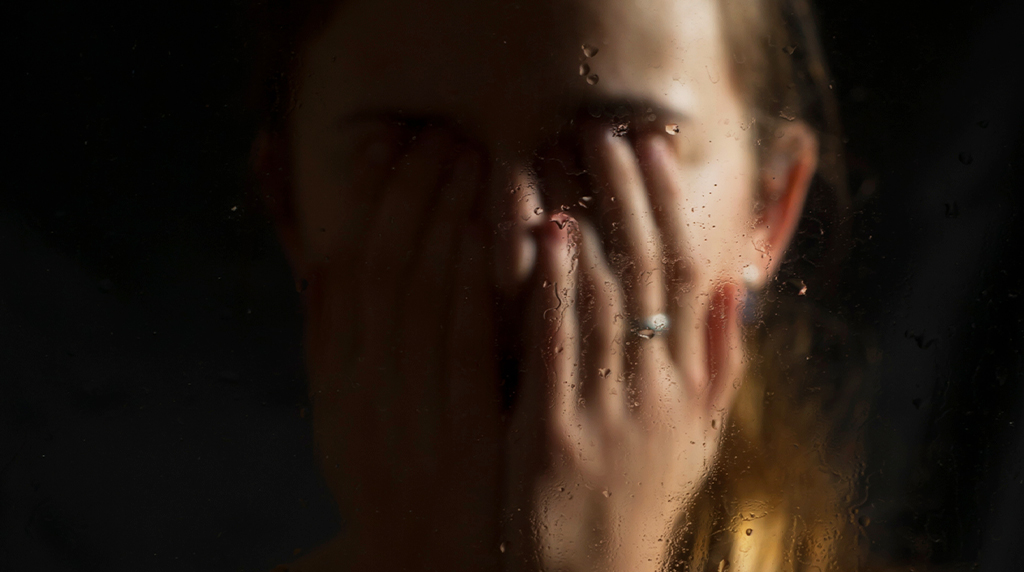 A person covers their face with both hands behind a rain-speckled glass conveying introspection or sadness.